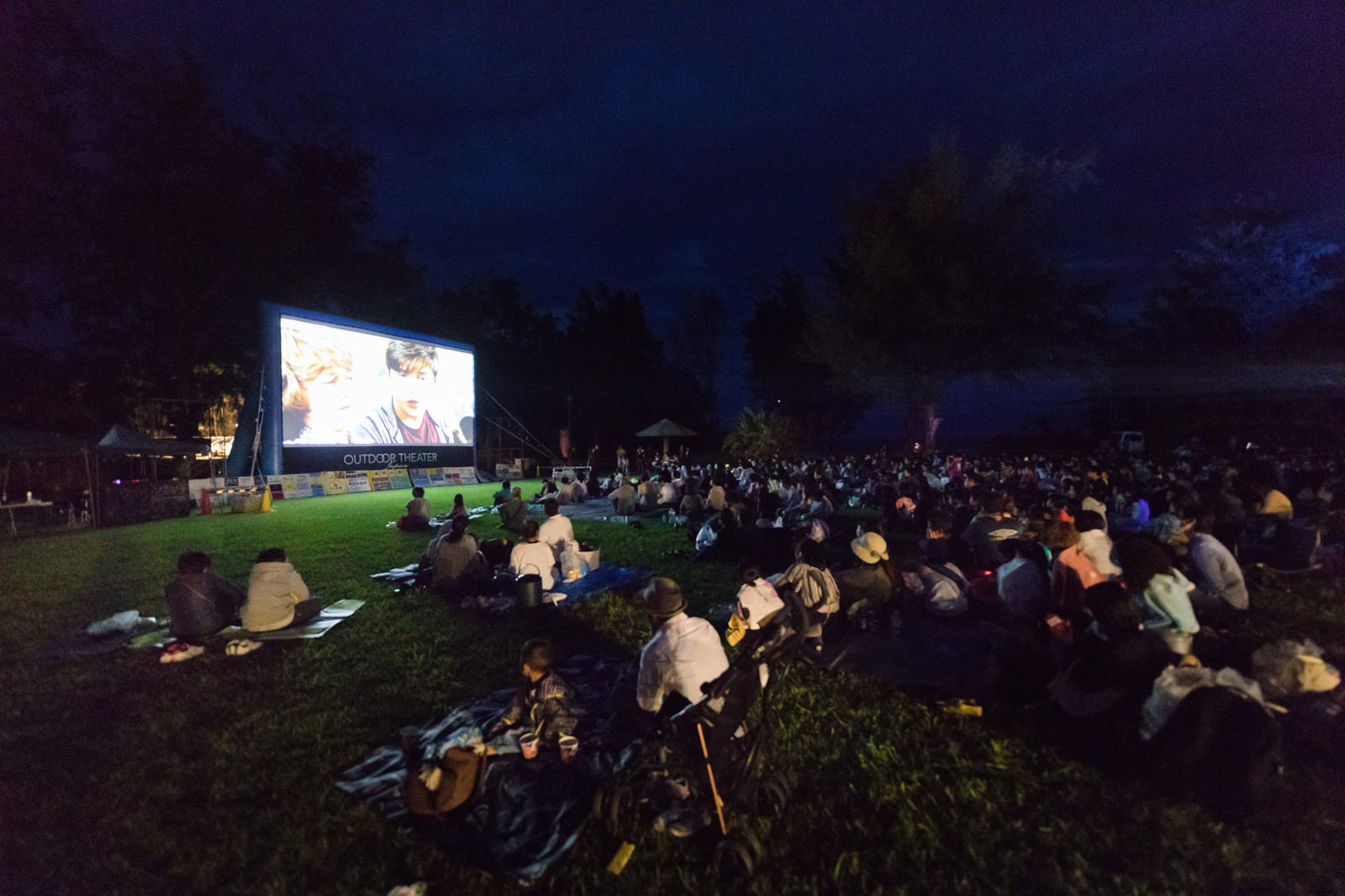 夜空の下、大きなスクリーンの前に座って映画を鑑賞する観客たち