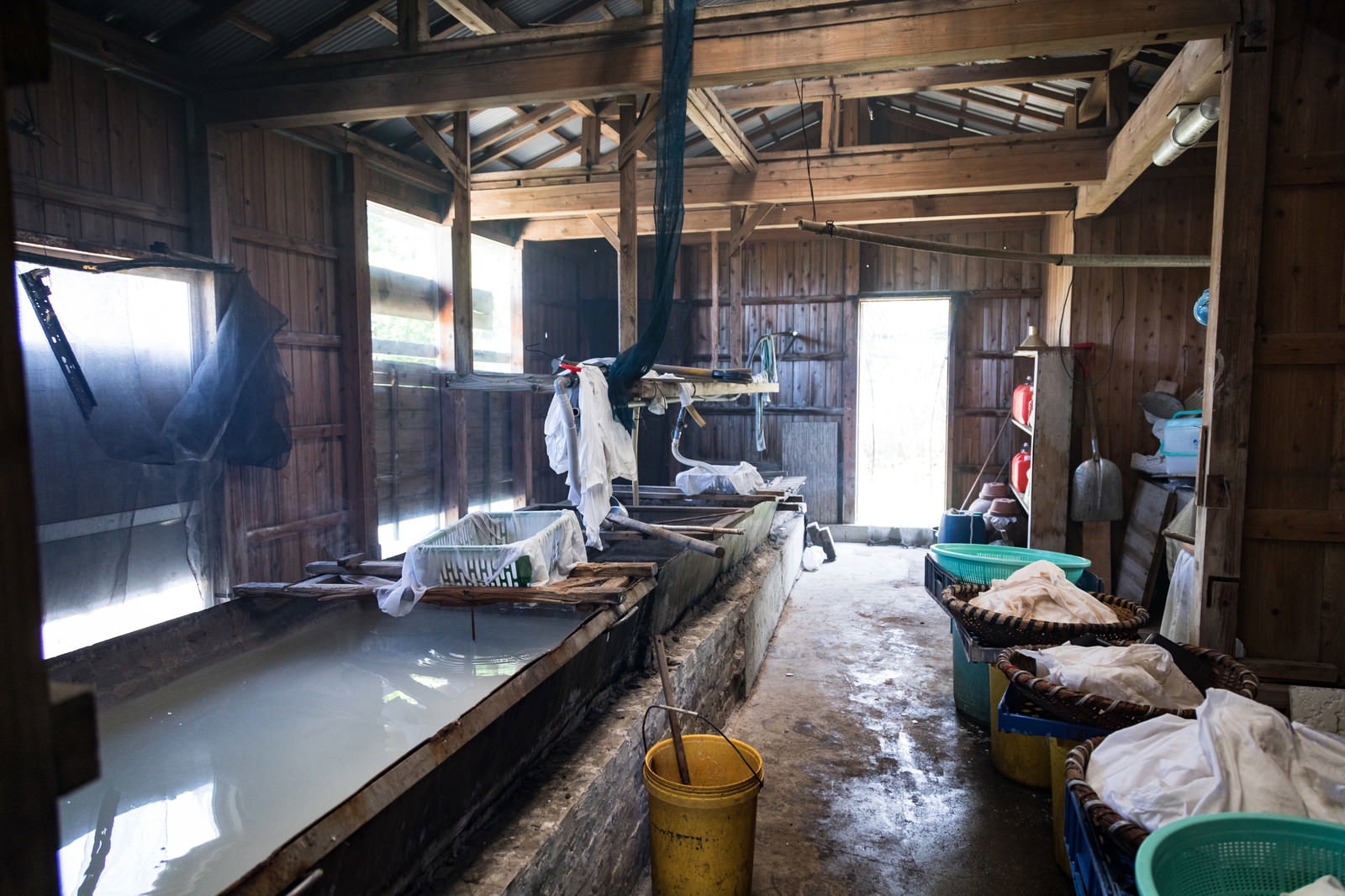 木造製塩小屋内の作業場。石造水槽、釜、かご、容器などが配置され、天井は梁と波板屋根で構成。