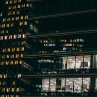 高層ビル群の窓ガラスから見える残業中のサラリーマンの写真