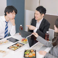 オフィスでランチミーティングしながら弁当を食べるビジネスパーソンの写真