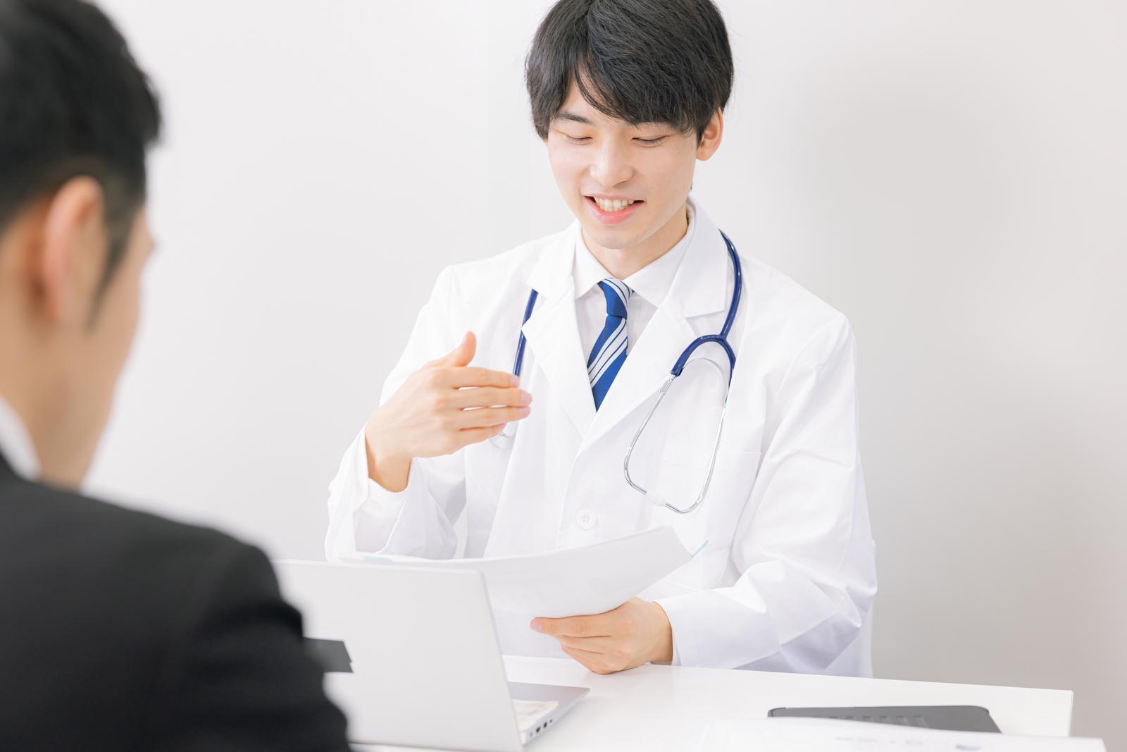 患者に医療アドバイスをする白衣の若い男性医師［モデル：たくみ］