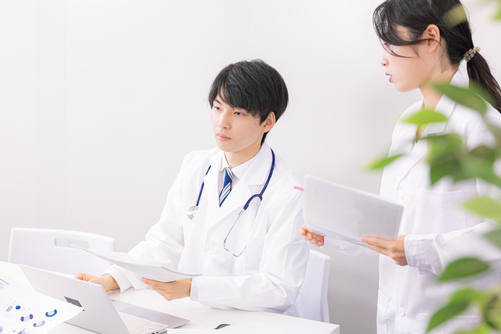 タブレットを見ながら打ち合わせをする白衣姿の男性医師と女性医師［モデル：たくみ 吉屋ゆり］