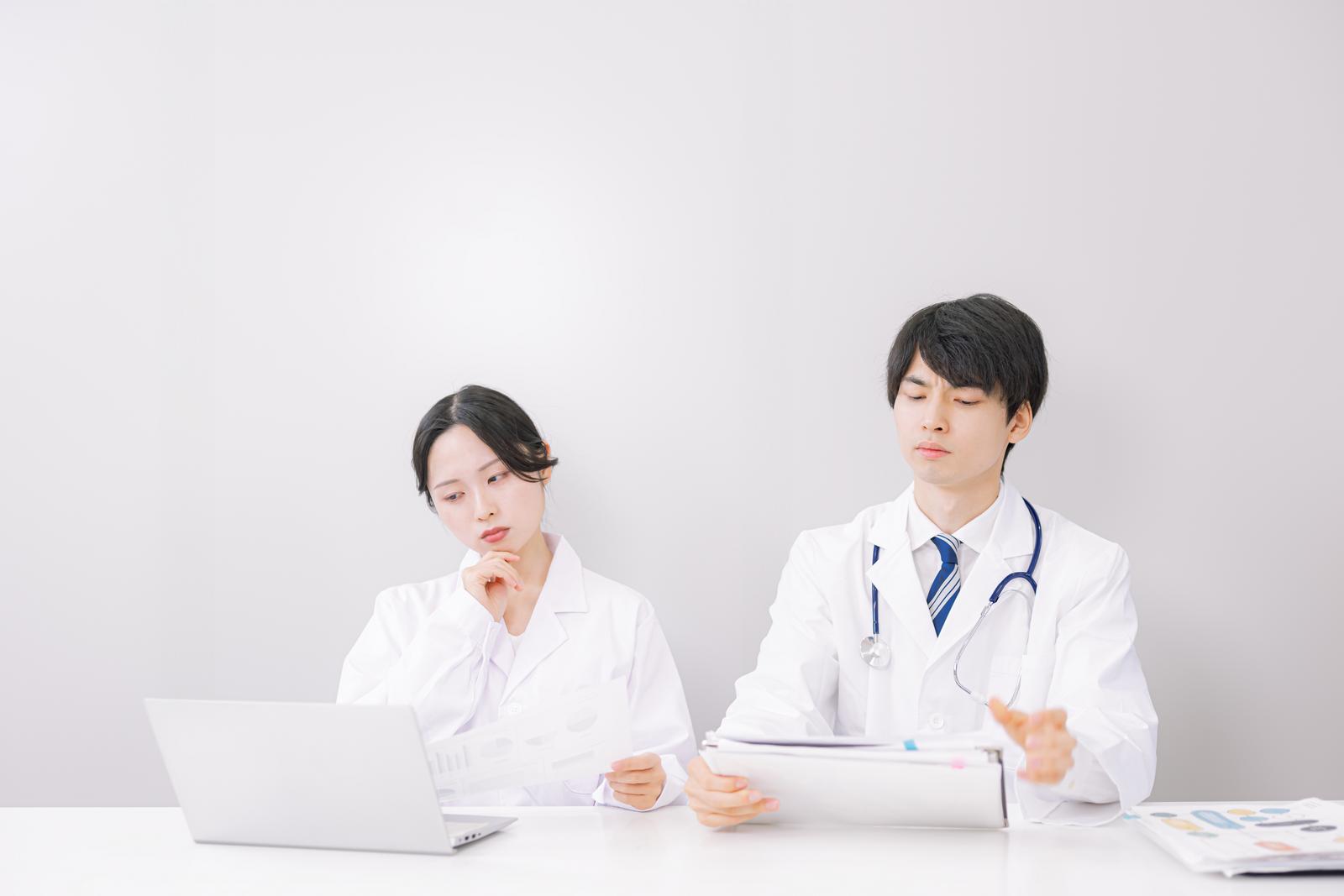 白衣の男性医師と女性医師がカルテとパソコンを確認している［モデル：たくみ 吉屋ゆり］
