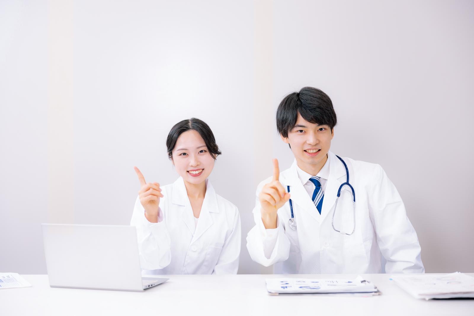 白衣を着た医師と研究員がデスク前で指を立てて笑う［モデル：たくみ 吉屋ゆり］