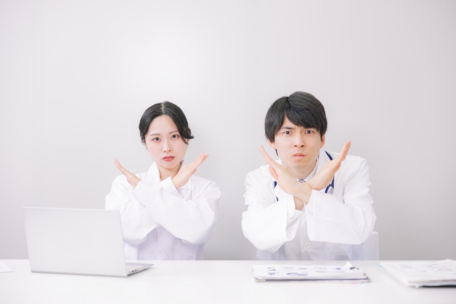 白衣を着た医師2人が机で禁止のジェスチャーをしている［モデル：たくみ 吉屋ゆり］