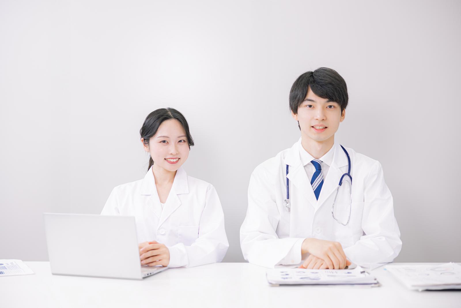 白衣を着たデスクの前で笑顔を見せる男性医師と女性医師［モデル：たくみ 吉屋ゆり］