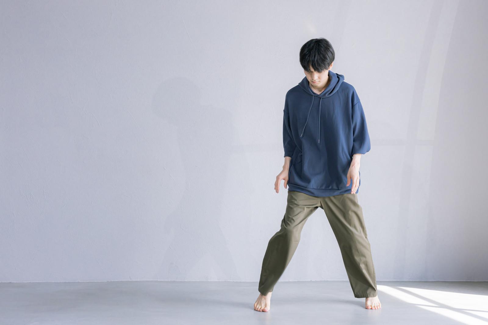 紺色のパーカーとカーキパンツを着た男性が、白い壁の前で裸足で立つポートレート写真［モデル：たくみ］