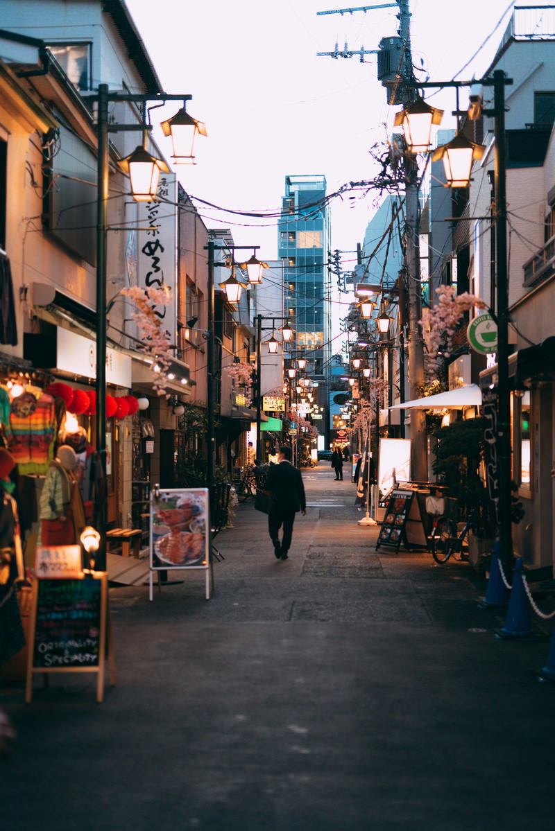 夕暮れ時の飲食店街で、両側に店が並ぶ狭い路地裏を一人で歩く通行人
