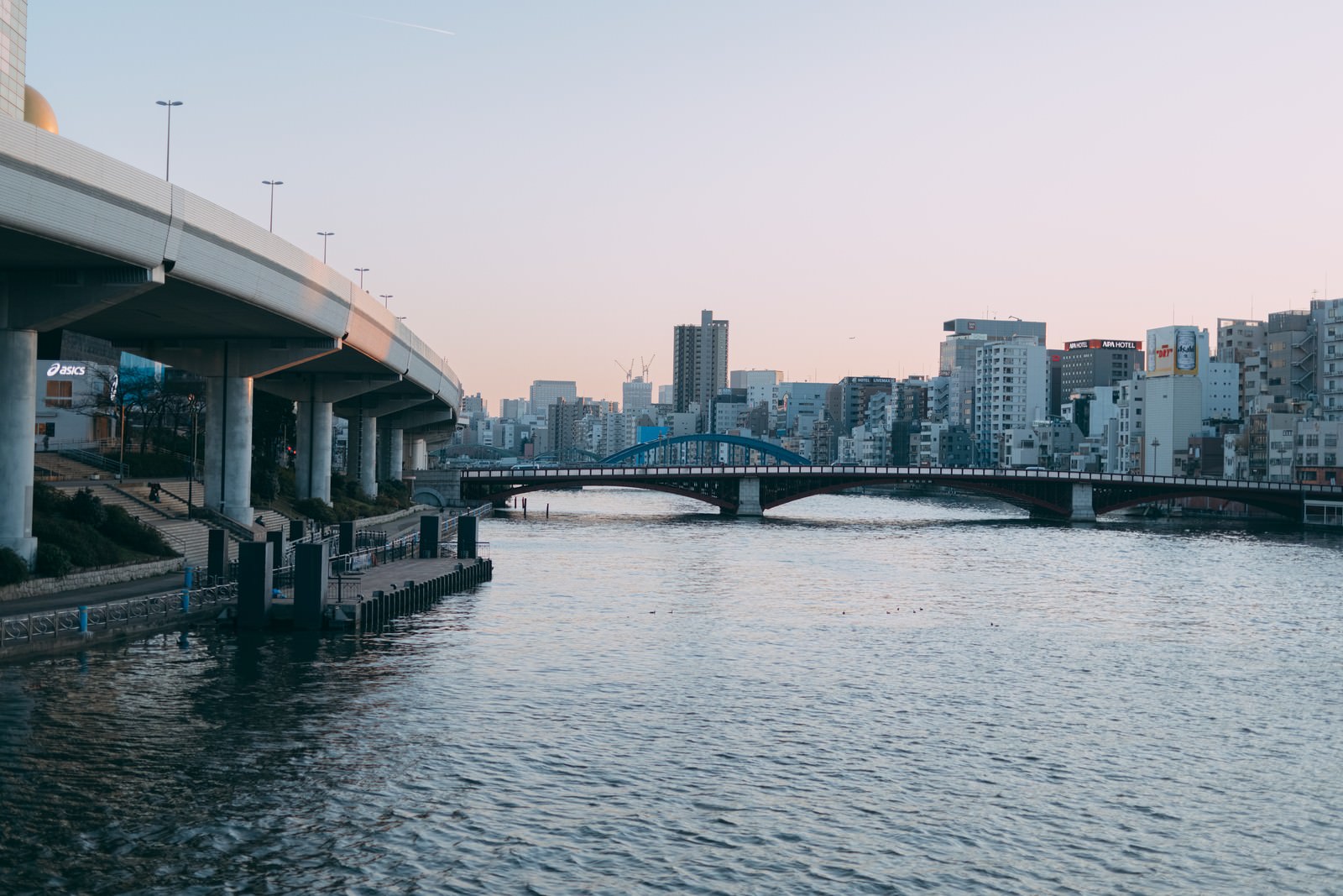夕暮れ時の隅田川に架かる橋と船着場、都市の建物群が見える風景