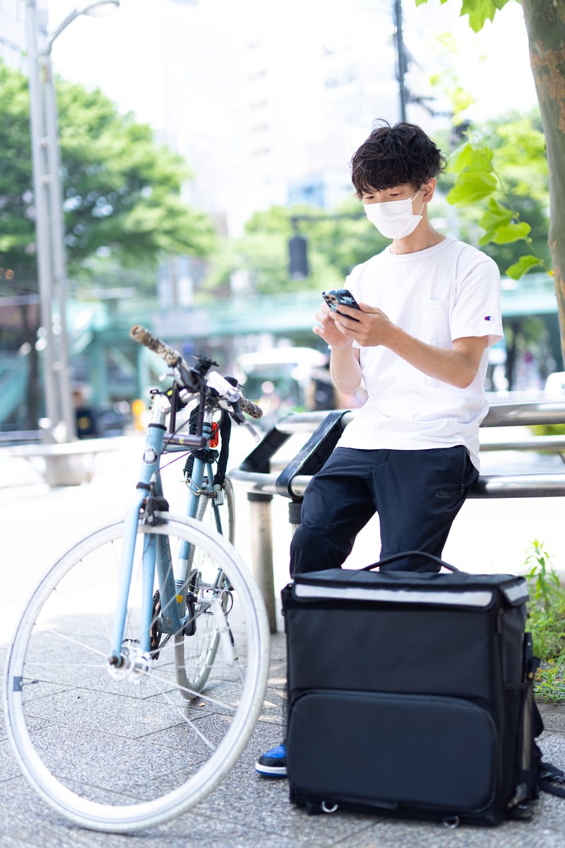 自転車の横でスマートフォンを見る白いTシャツの配達員男性［モデル：TSURUTAMA］
