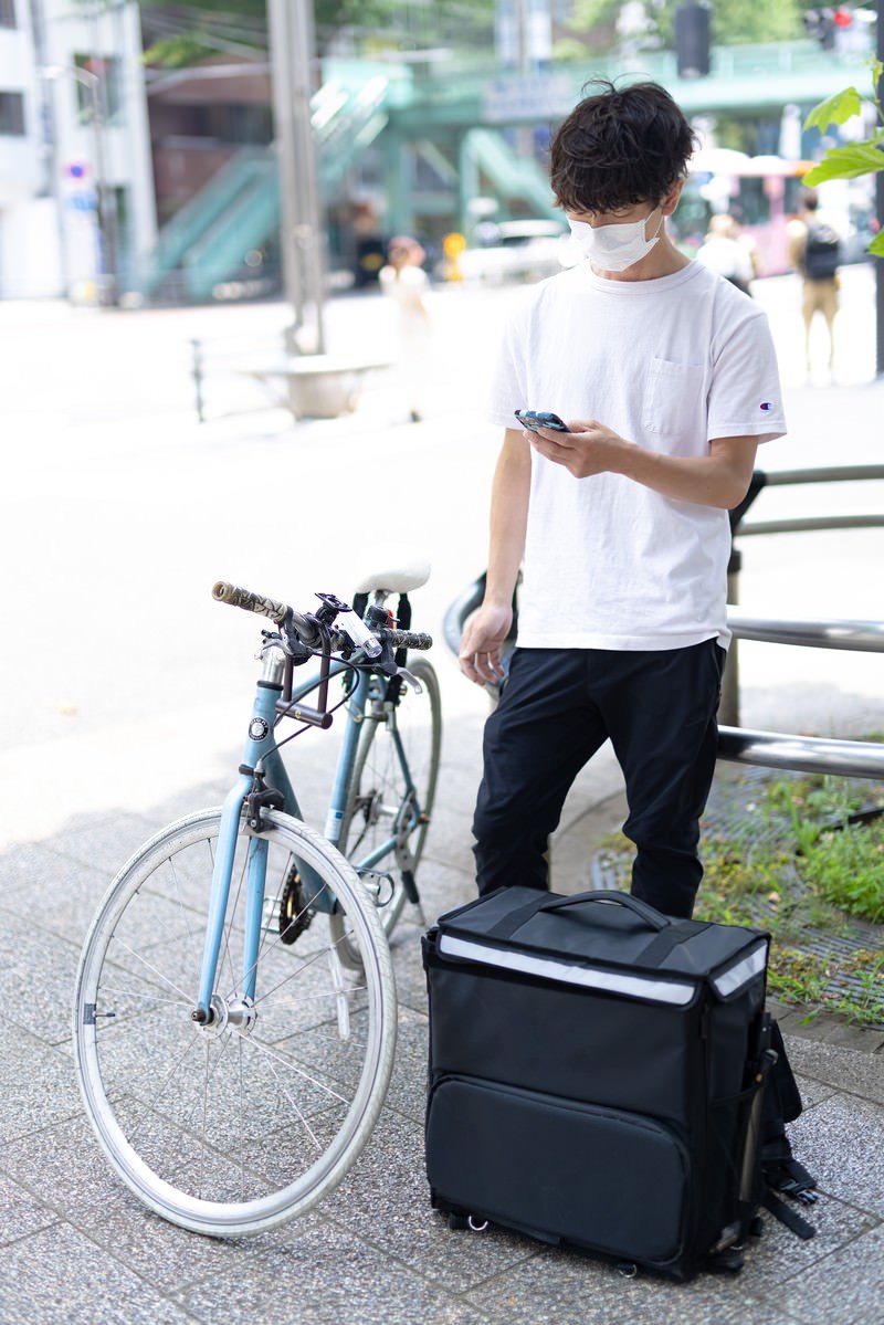 宅配用の黒いリュックを背負い自転車の横に立つ配達パートナーの男性［モデル：TSURUTAMA］