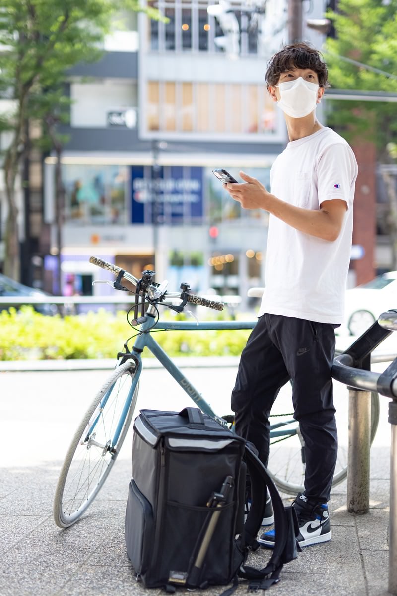 配達先を探すため自転車の横でスマートフォンを見る白いTシャツの配達員男性［モデル：TSURUTAMA］