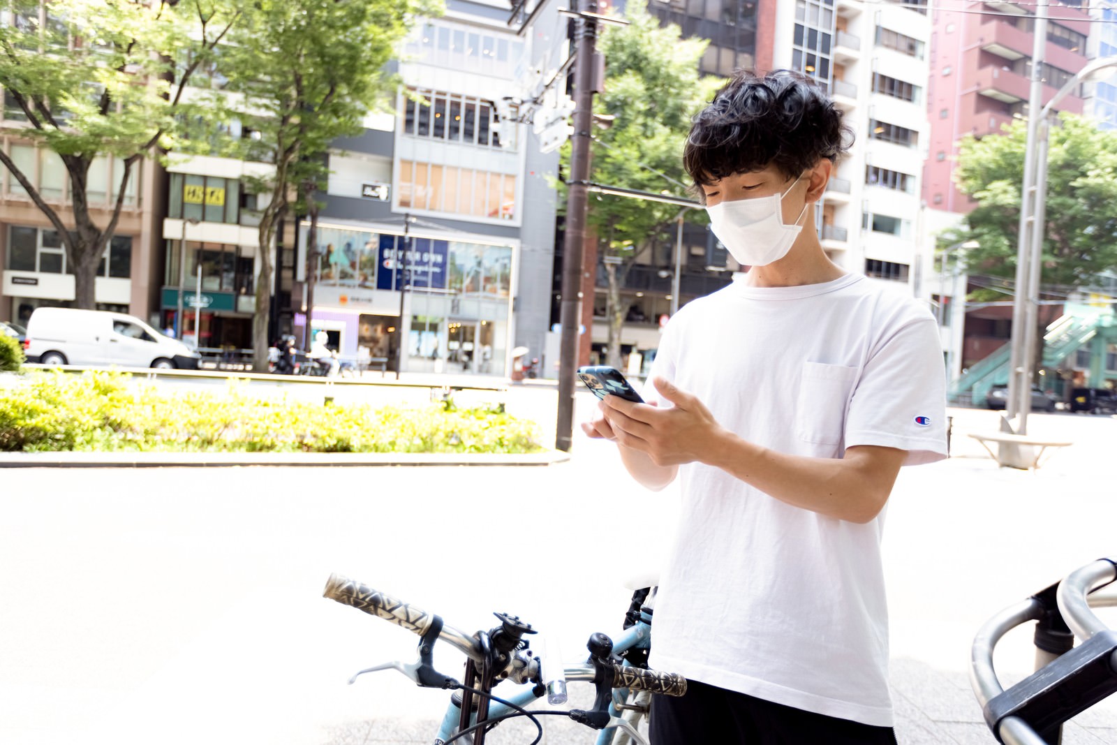 自転車の前でスマートフォンを操作する白いTシャツの男性配達員［モデル：TSURUTAMA］