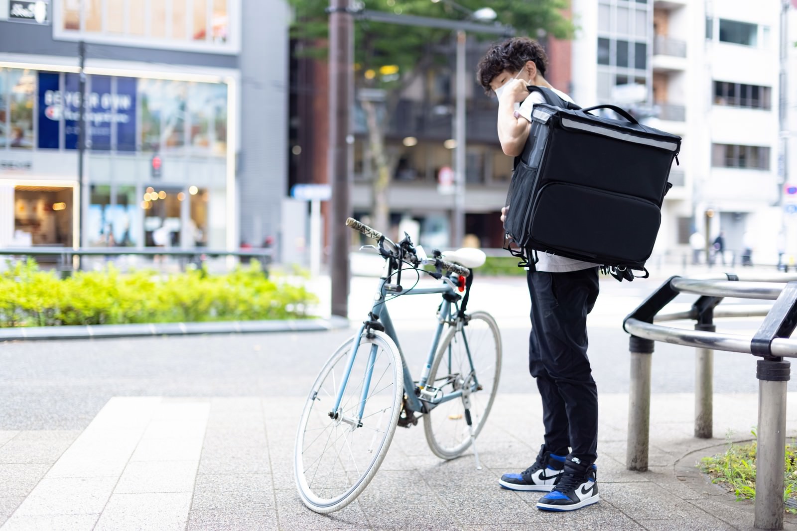 ウーバーイーツのデリバリーバッグを背負い自転車の前に立つ男性配達員［モデル：TSURUTAMA］