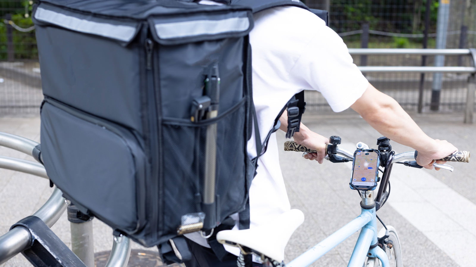 黒い配達バッグを背負い、スマートフォンの地図を見ながら自転車で移動する配達員の後ろ姿［モデル：TSURUTAMA］