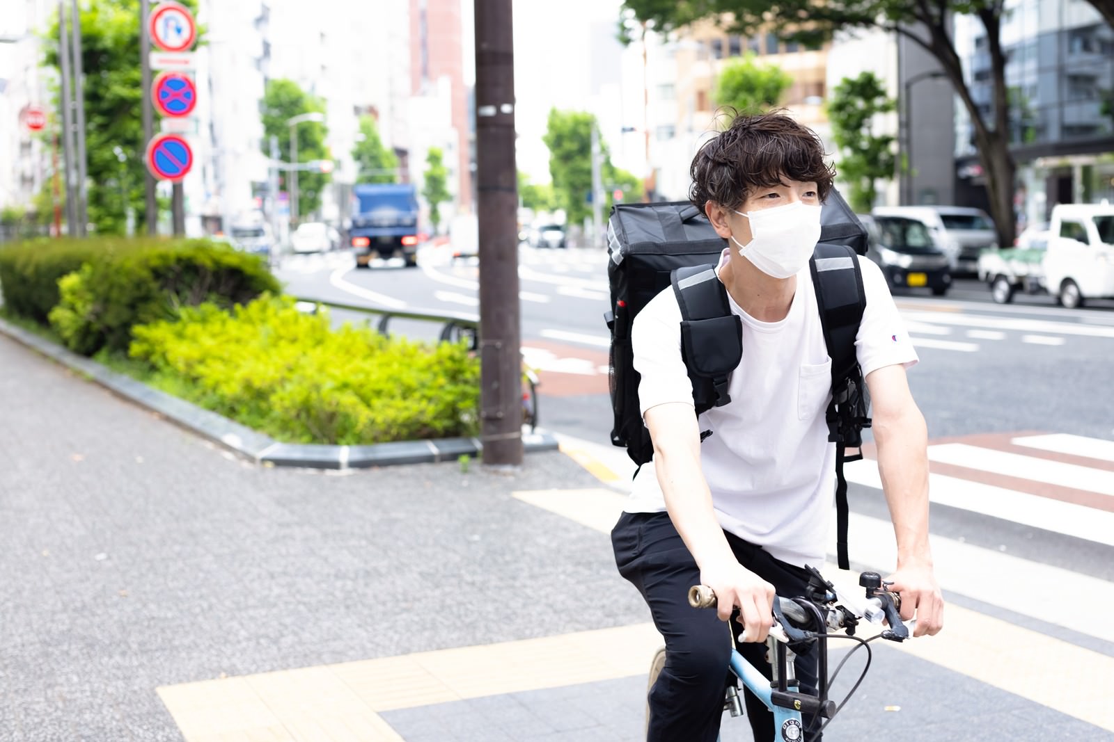 白いヘルメットを被りリュックを背負った配達員が自転車で信号待ちをしている［モデル：TSURUTAMA］