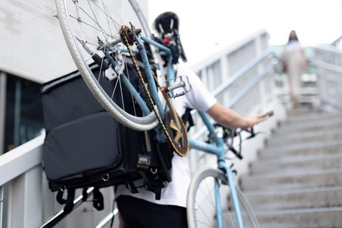 歩道橋の階段を自転車を背負って登る配達員の男性