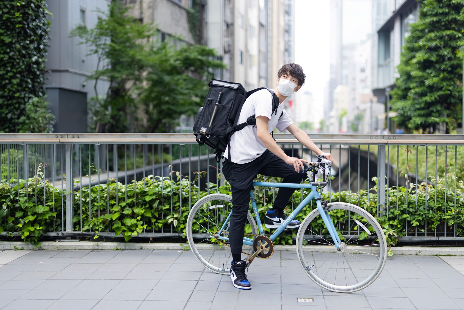 黒いリュックを背負った配達員の男性が自転車と一緒に歩道橋に立っている［モデル：TSURUTAMA］