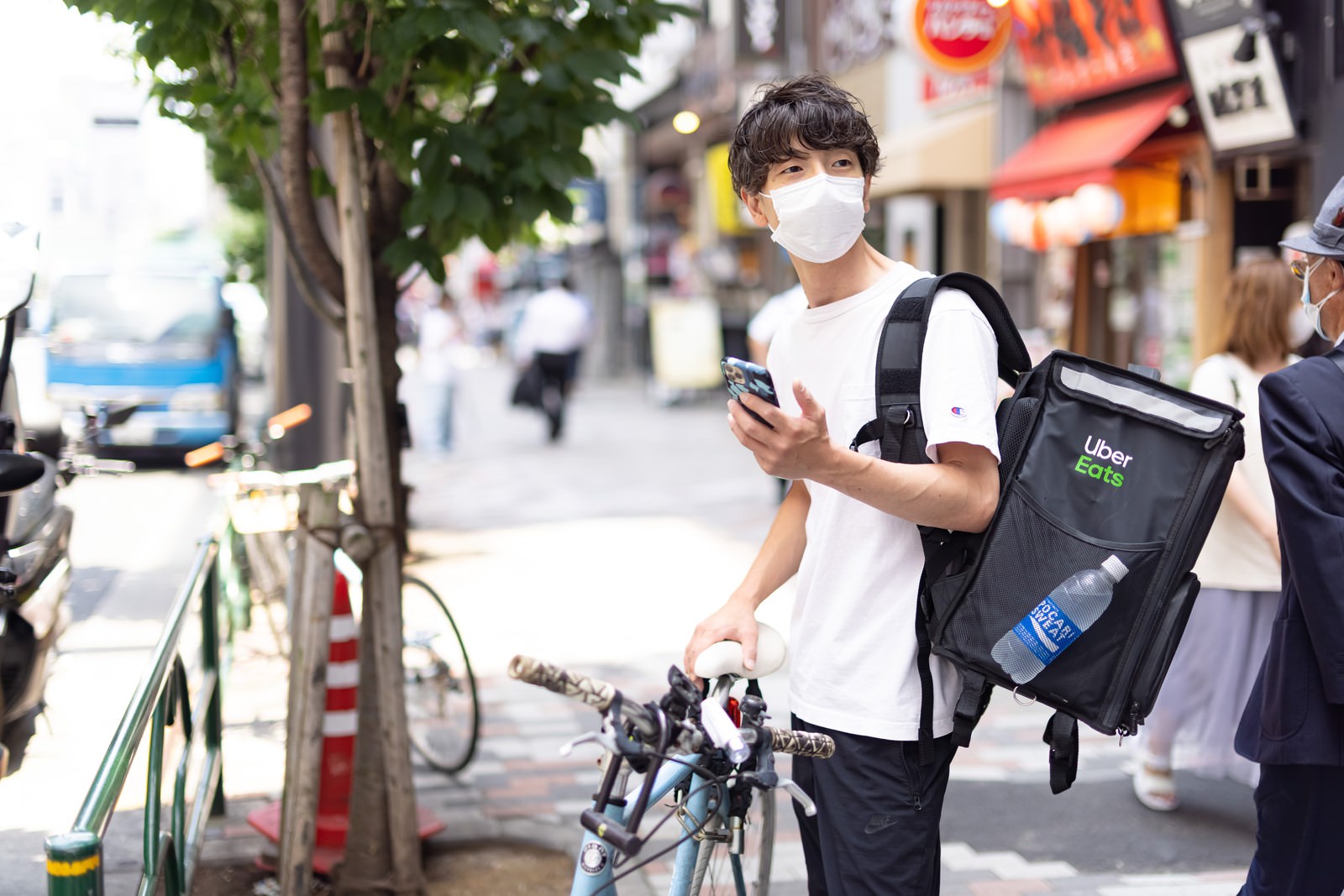 マスクをつけた配達員が黒いリュックを背負って自転車の横に立っている様子［モデル：TSURUTAMA］