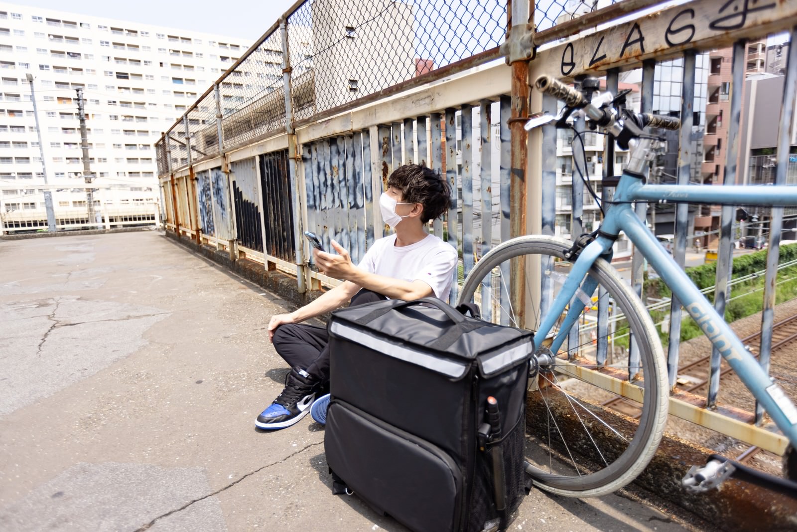 配達バッグを背負った男性配達員が自転車の横でスマートフォンを見ながら休憩している［モデル：TSURUTAMA］