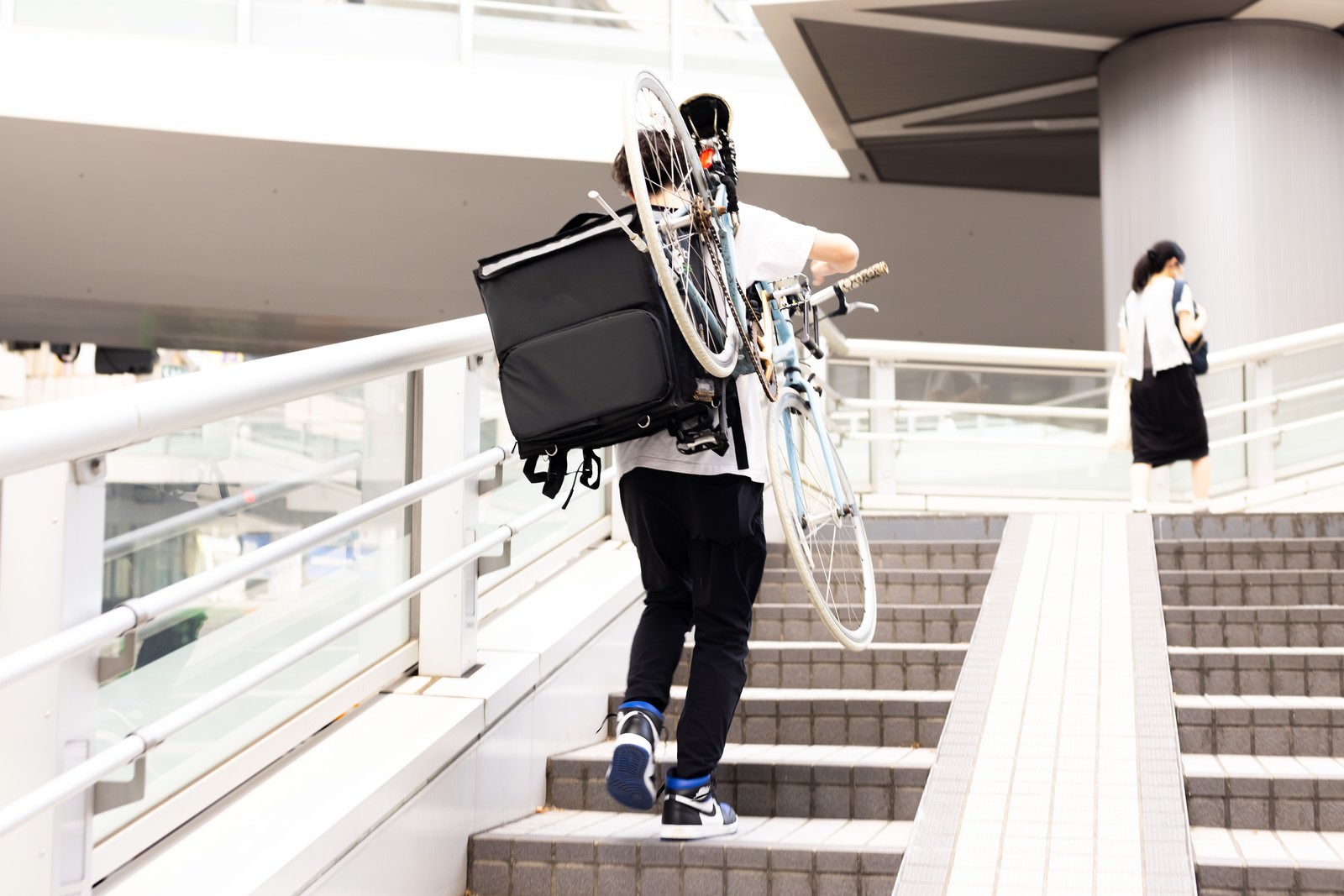 黒い服装の配達員が白い階段で自転車を担いで上っている様子［モデル：TSURUTAMA］
