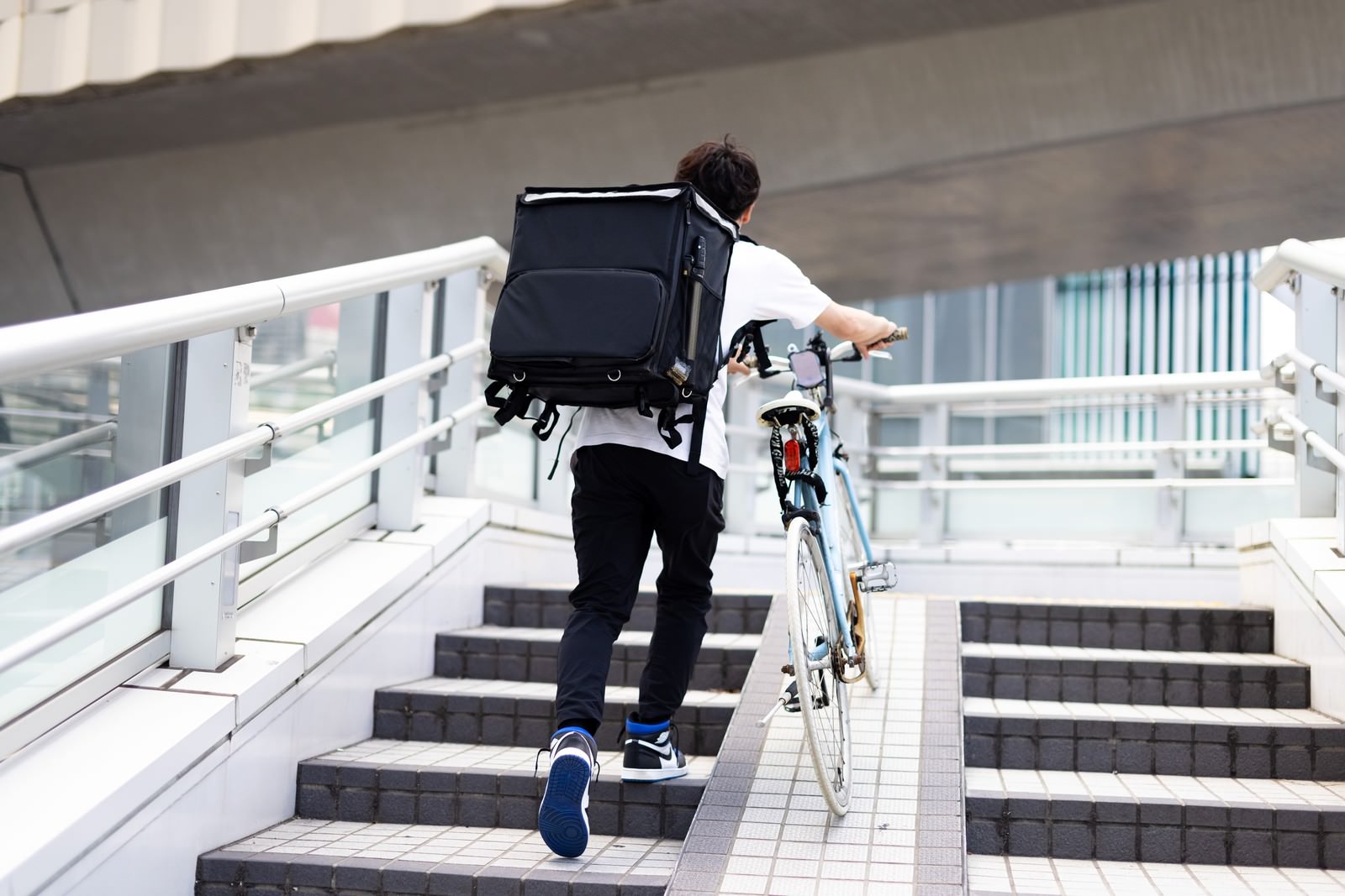 黒いデリバリーバッグを背負い自転車を押して階段を上る配達員［モデル：TSURUTAMA］