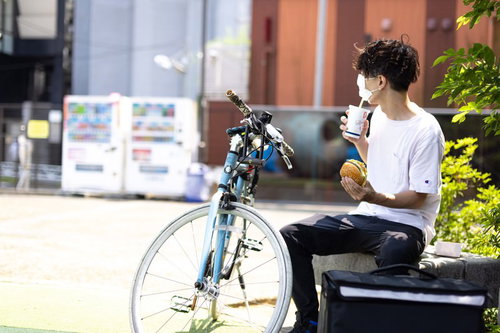フードデリバリーの配達員が自転車の前で休憩中にハンバーガーを食べている