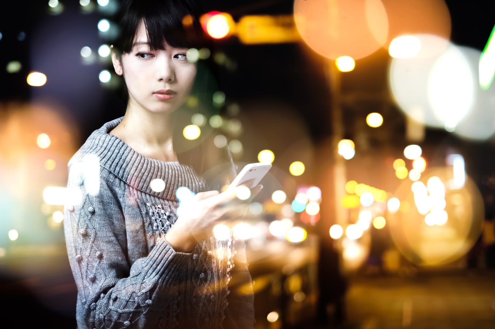 スマートフォンを持ちながら、ぼやけた夜景の街を背景に立つ女性［モデル：白鳥片栗粉］
