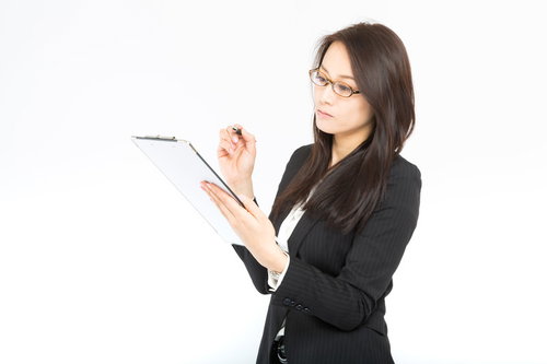 クリップボードとペンを持ちチェックする眼鏡の黒スーツの長い黒髪の女性
