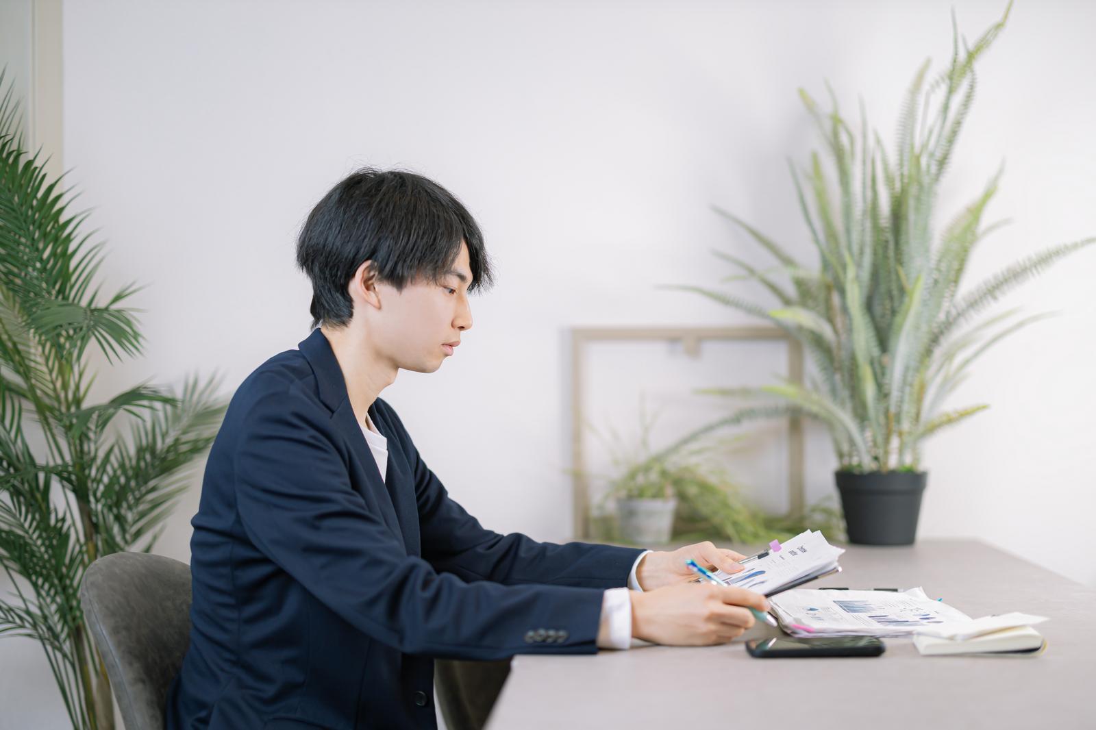 デスクで書類を見ながらペンで記入するビジネスパーソン［モデル：たくみ］
