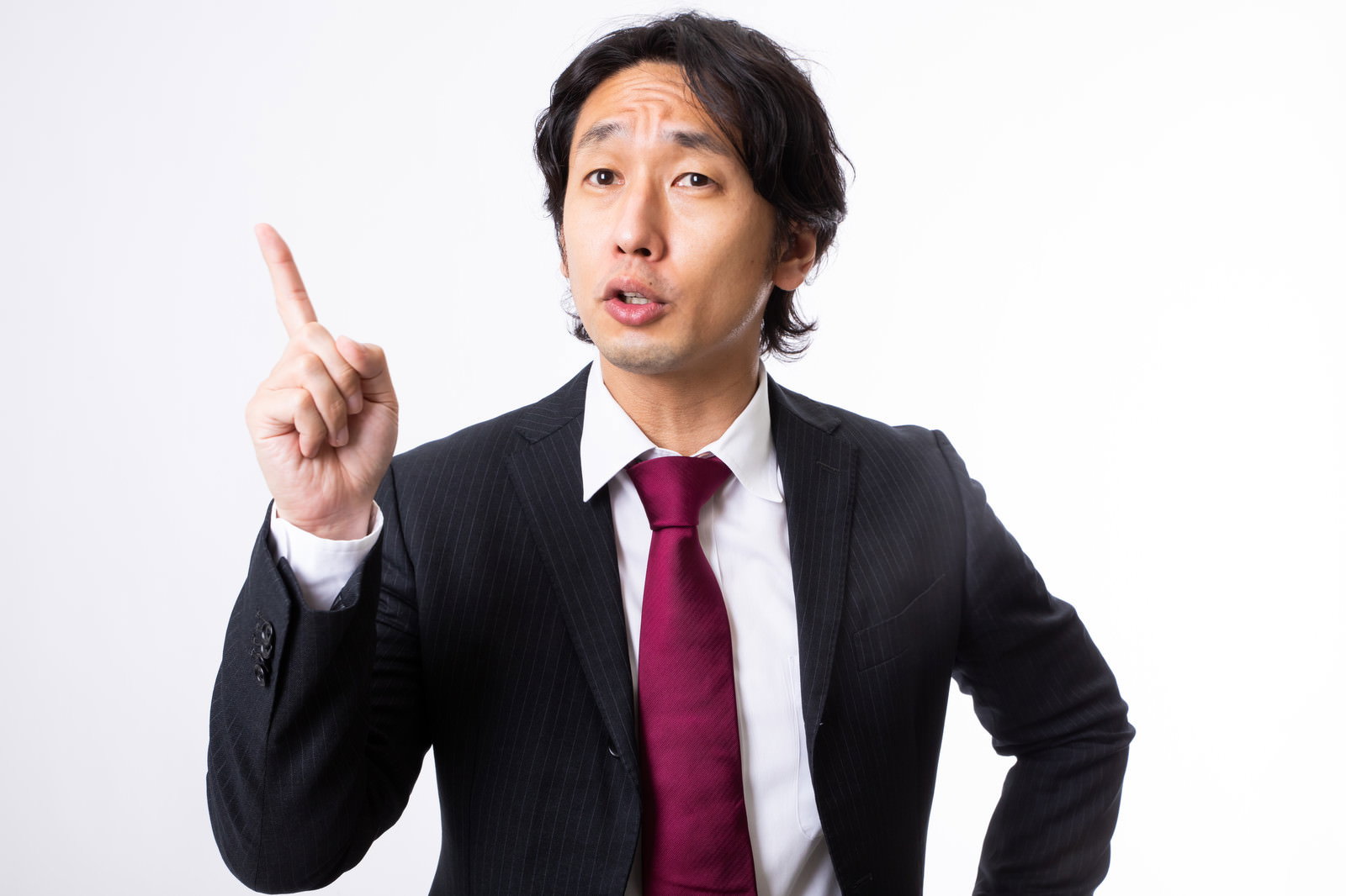 黒いスーツに赤いネクタイの男性が右手の人差し指を上に立てて指摘している［モデル：大川竜弥］