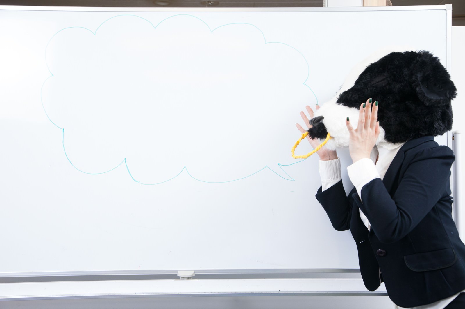 黒いスーツを着た牛がマーカーペンでホワイトボードに吹き出しを描き、背後に人物が見守っている［モデル：古性のっち］