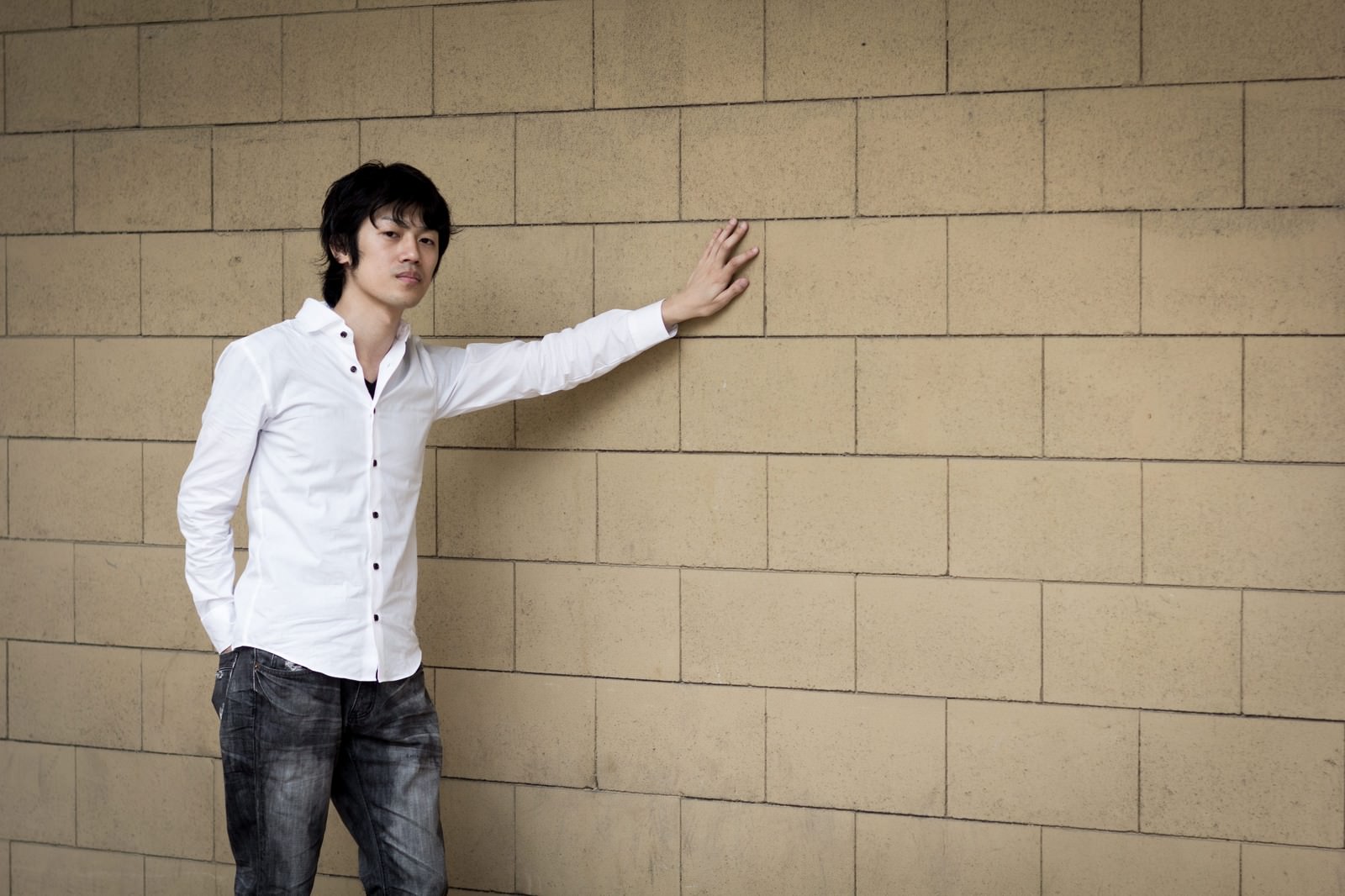 白シャツを着た男性が壁の前で片腕を伸ばすポーズをしている［モデル：Tsuyoshi.］