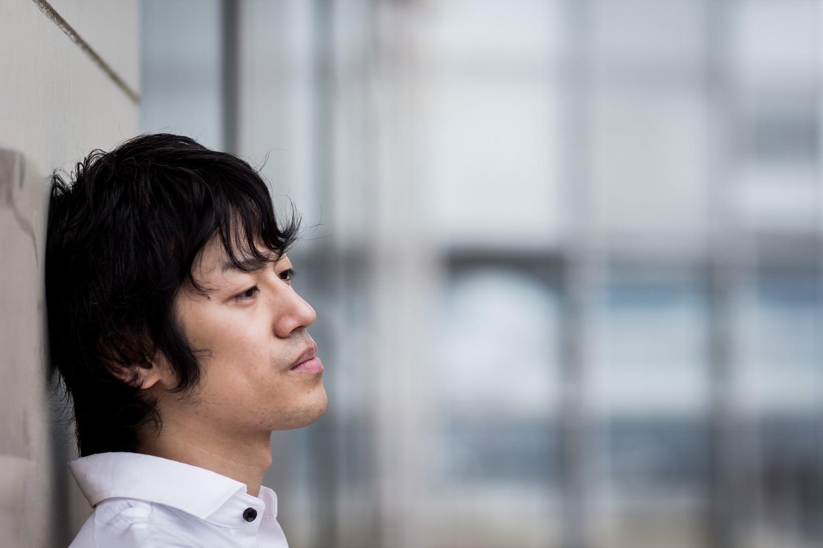 壁にもたれかかり下を向く白いシャツの男性［モデル：Tsuyoshi.］