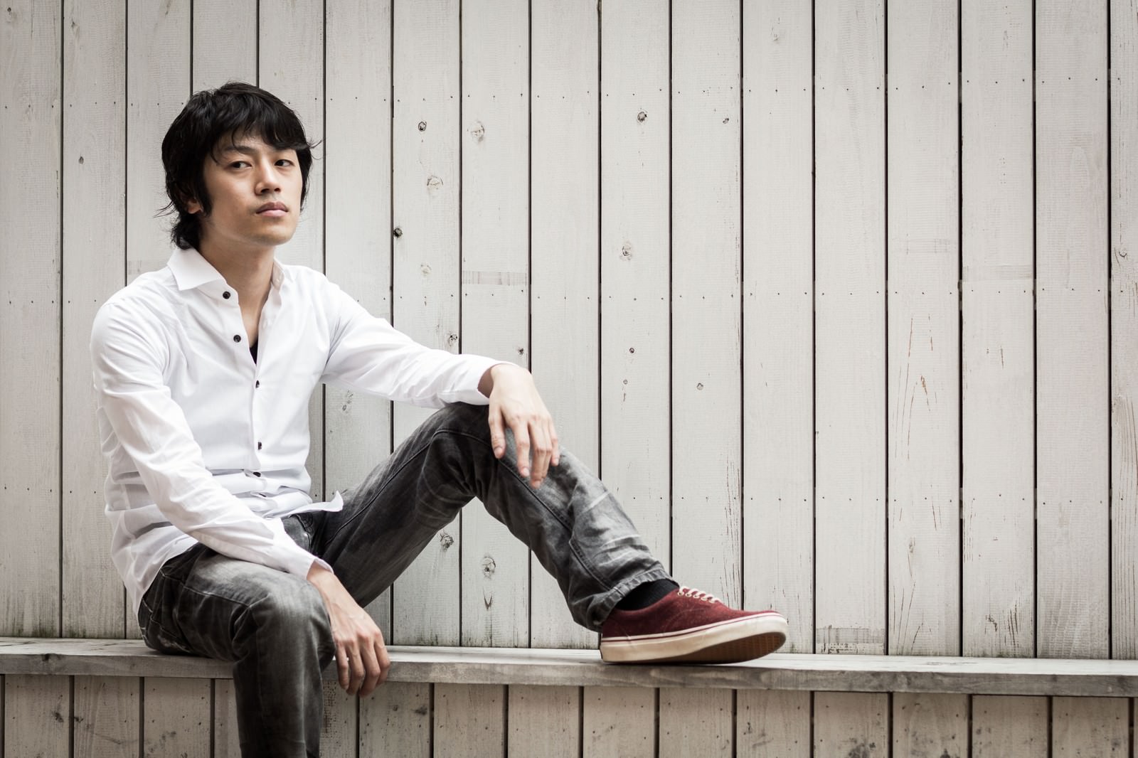 白シャツを着た男性が木製の壁を背景に座り込んでいる［モデル：Tsuyoshi.］