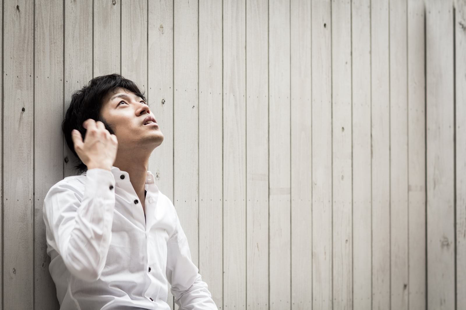 白いシャツを着た男性が壁を背に上を見上げている［モデル：Tsuyoshi.］