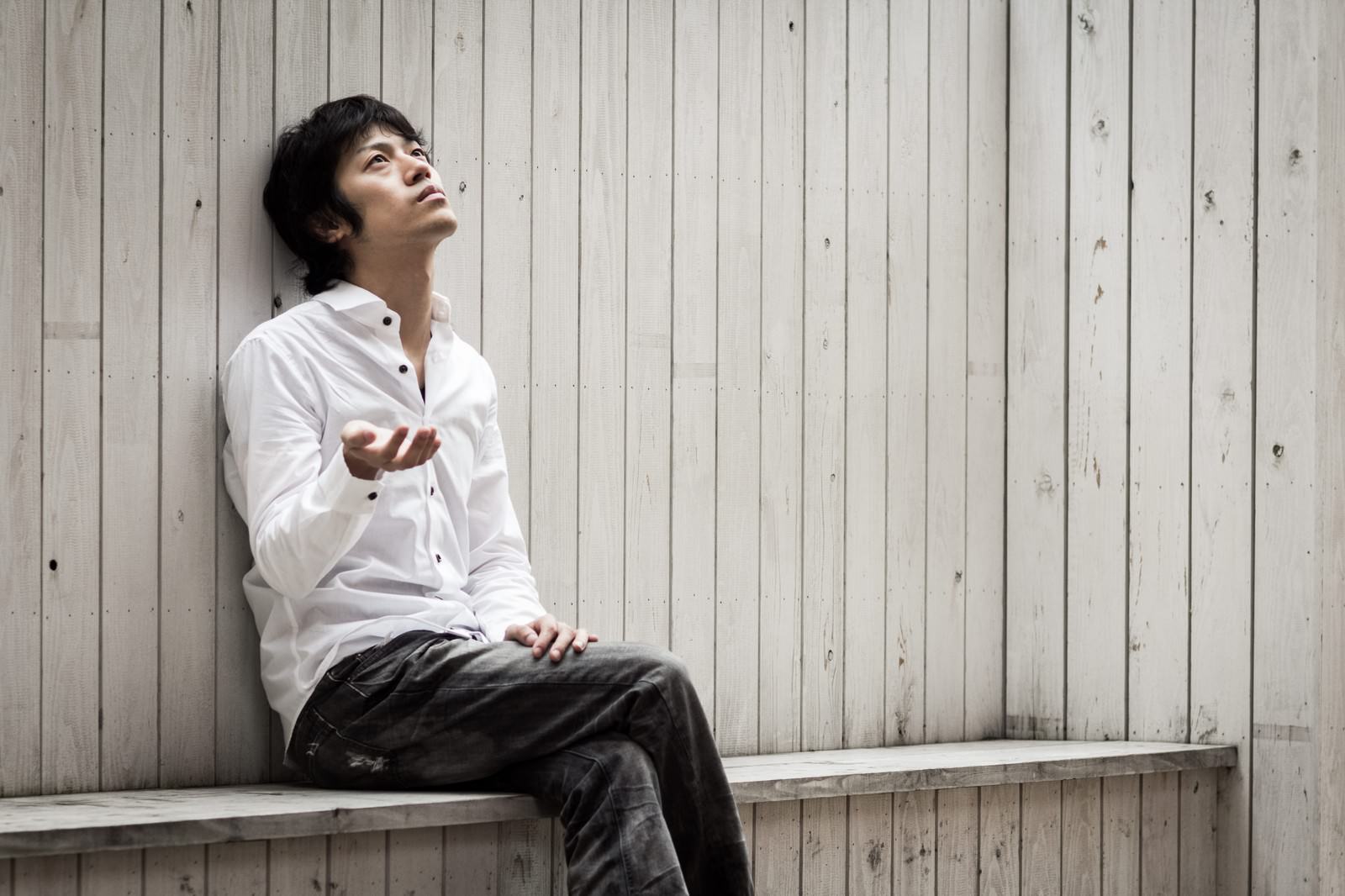 白いシャツを着た男性が木のベンチに座り、上を向きながら手をかざしている［モデル：Tsuyoshi.］