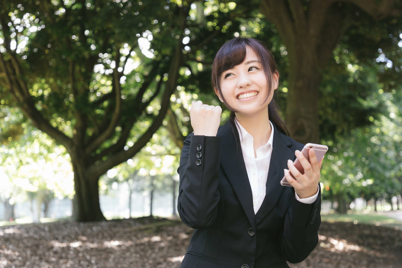スマートフォンを持ちながらガッツポーズをする黒スーツ姿の就活女性［モデル：河村友歌］
