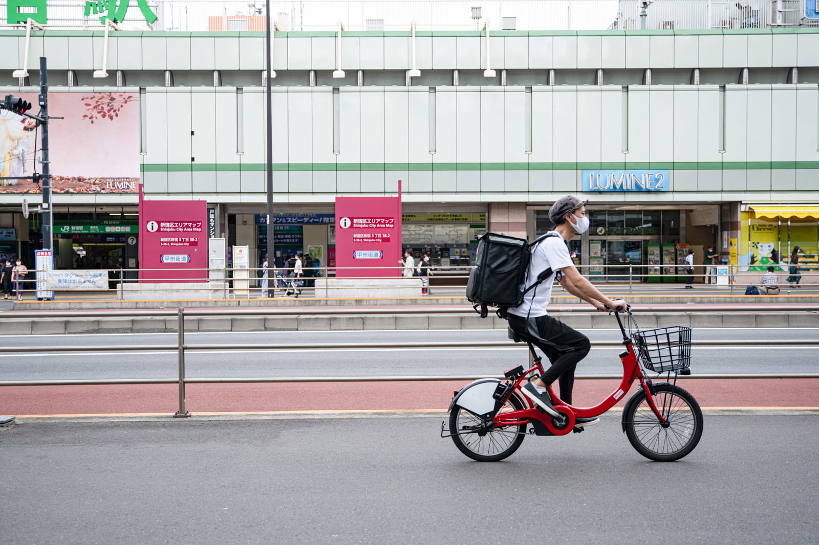 新宿駅前の道路を赤い自転車で走行するフードデリバリー配達員