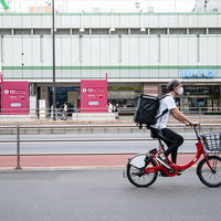 新宿駅前を赤い自転車で走行するフードデリバリー配達員の写真