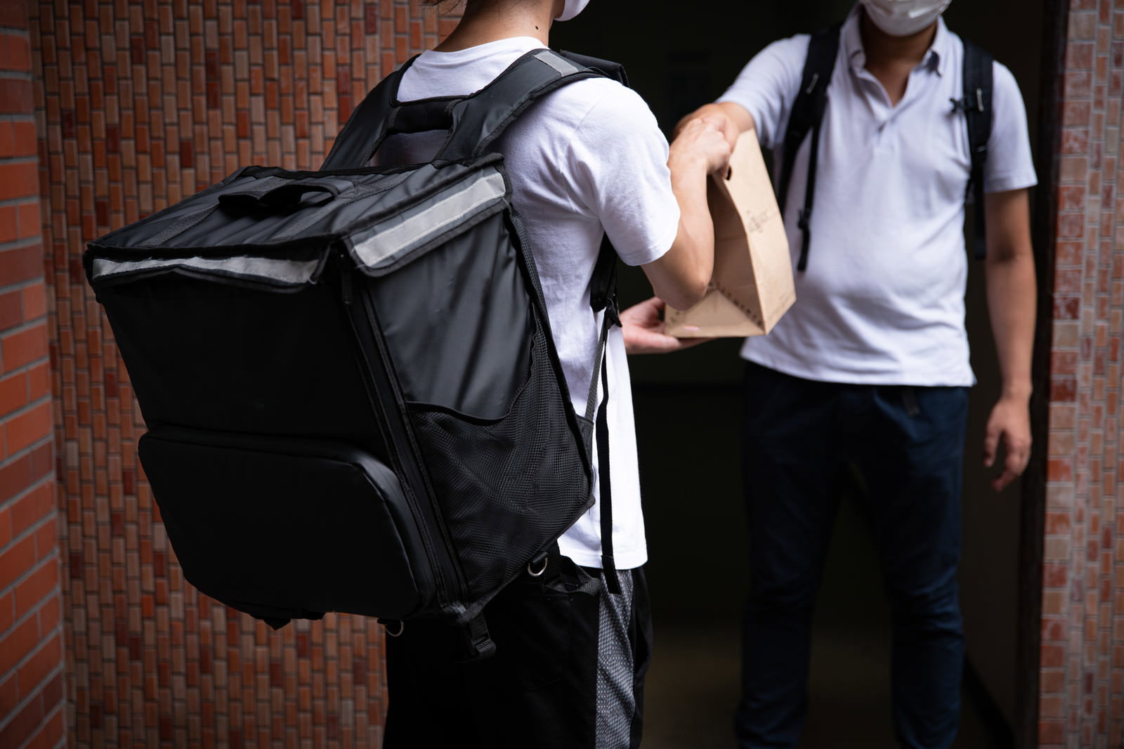 黒いデリバリーバッグを背負った男性が紙袋を手に持ち、受け取り人と対面して配達メモを確認している場面