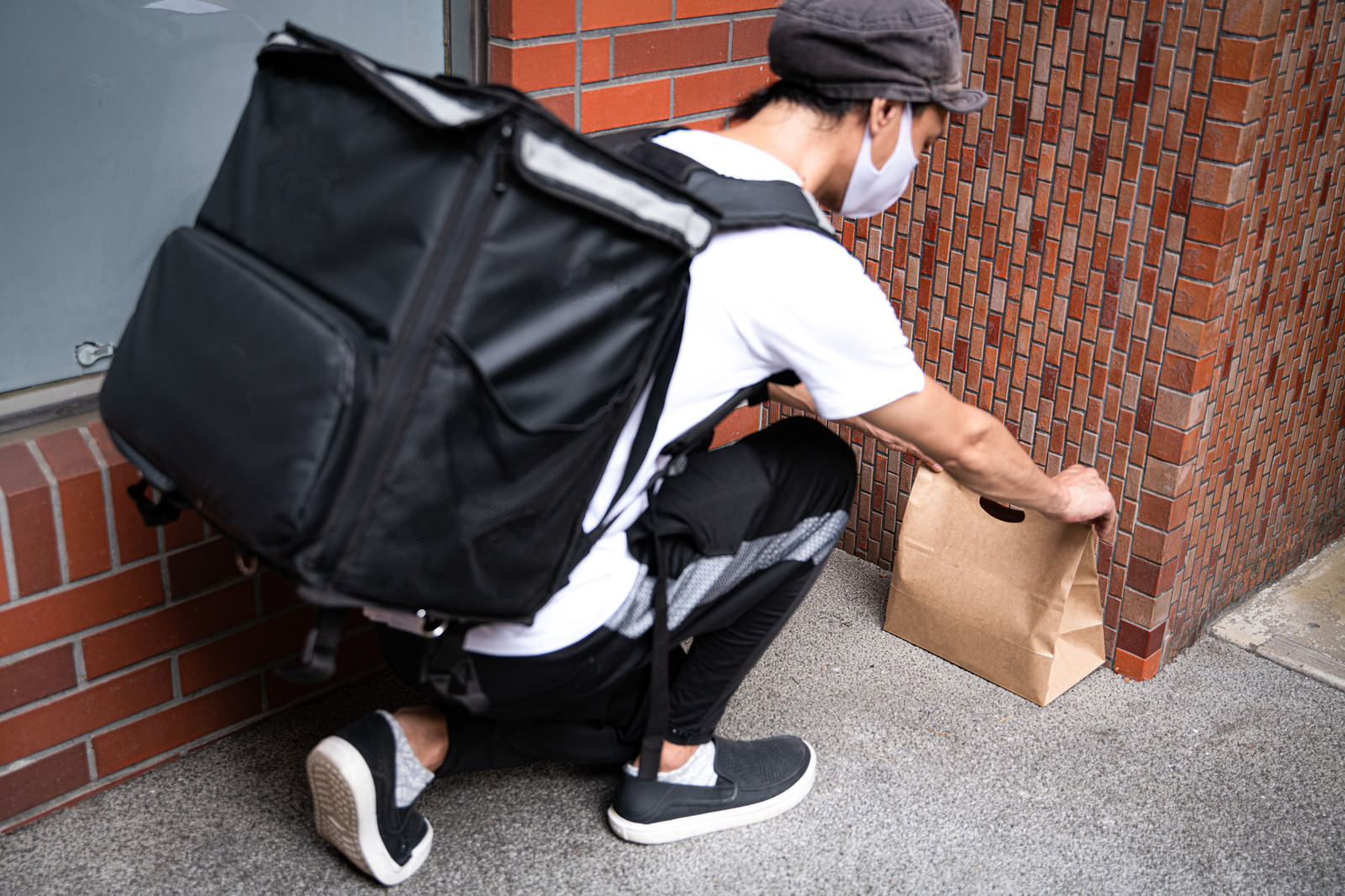 黒い配達バッグを背負い玄関先に紙袋を置き配する配達員
