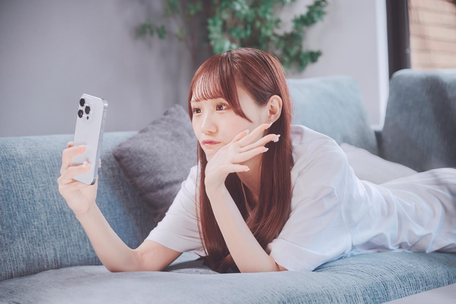 ソファーに寝転んでスマートフォンで自撮りする白いシャツの女性［モデル：あき］