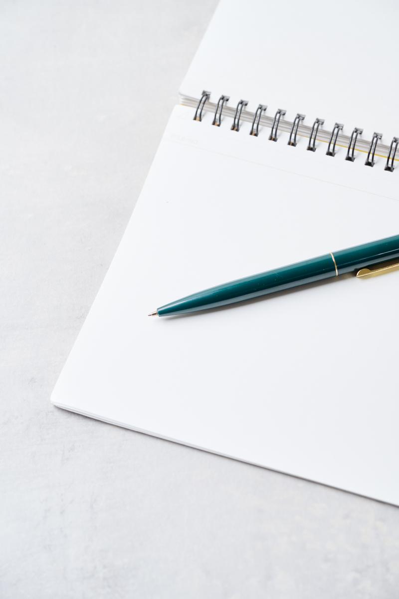 開いたスパイラルノートの横に置かれた緑色のボールペン