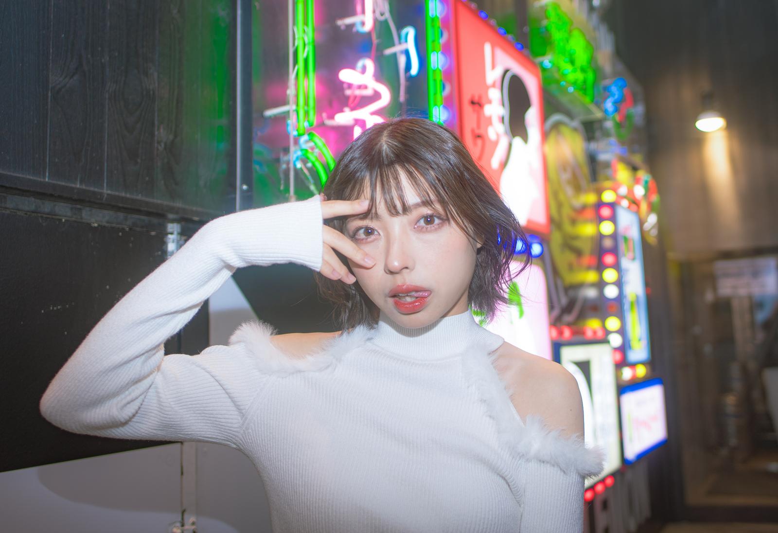 ネオン看板が光る飲み屋街で、白いタートルネックを着た女性が指を顔に当てるポーズ［モデル：高尾実生］