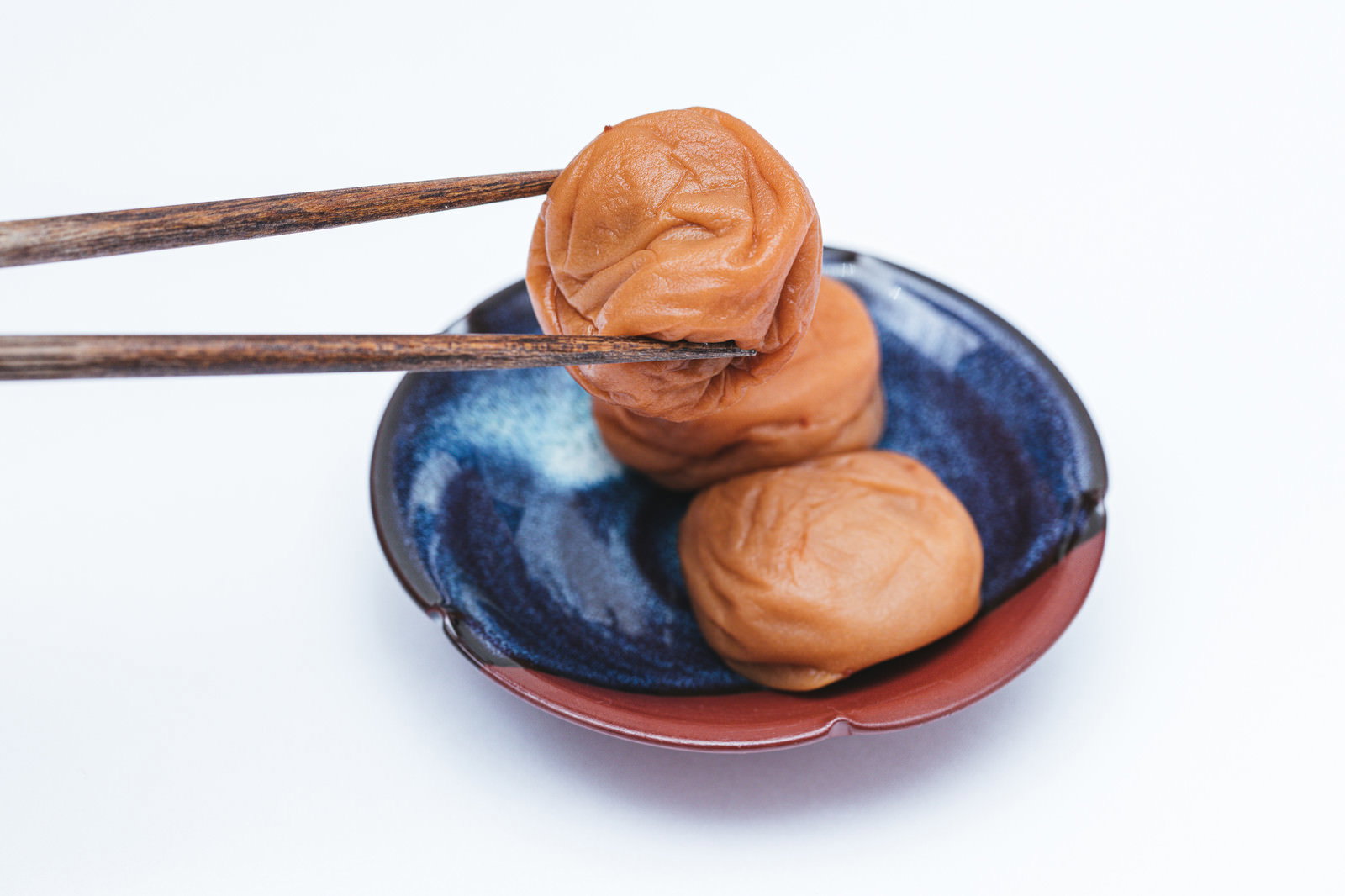 青い皿に盛られた梅干しを木製の箸で掴んでいる様子