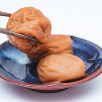 青い小皿の梅干しを箸でひとつつまむ瞬間の和食の写真