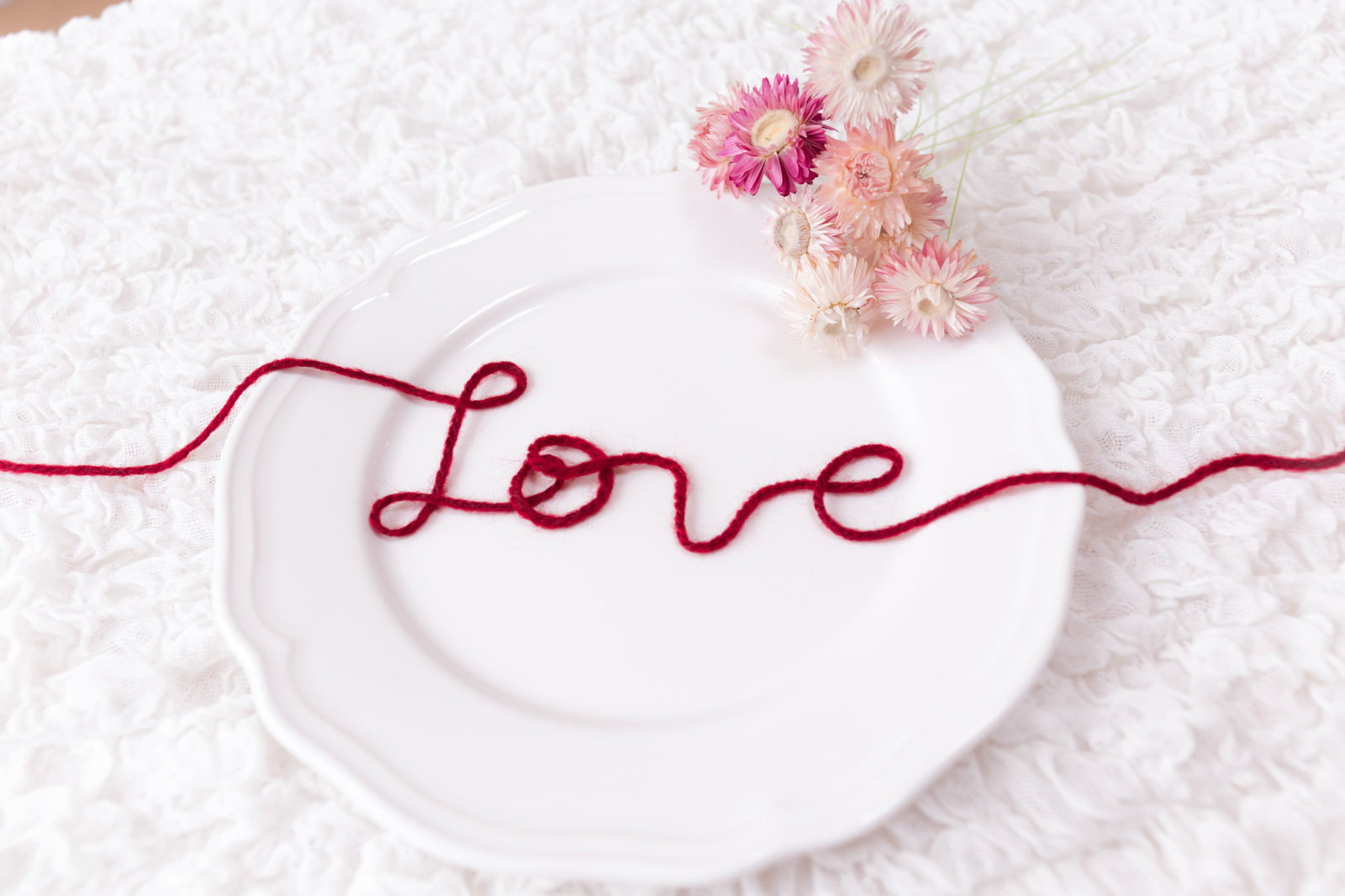 白い皿の上に赤い糸でLoveの文字を作り、ピンクの花を背景に配置した愛をテーマにした写真