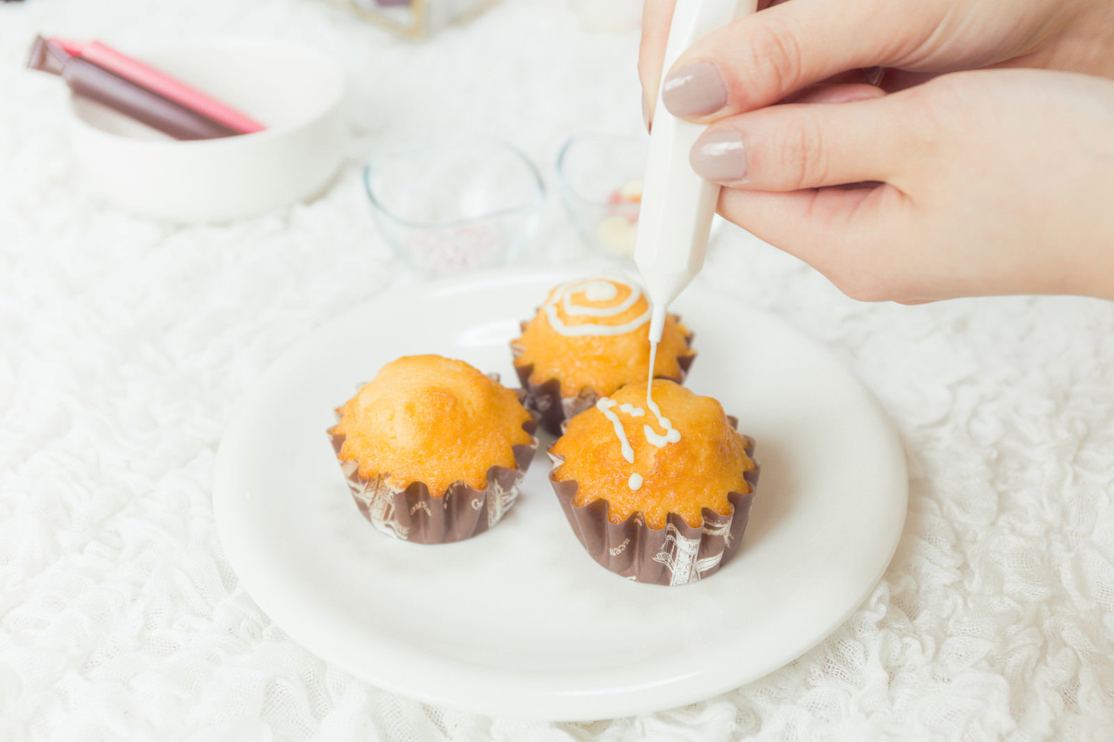 白い皿に並んだカップケーキにホワイトチョコをデコレーションする手