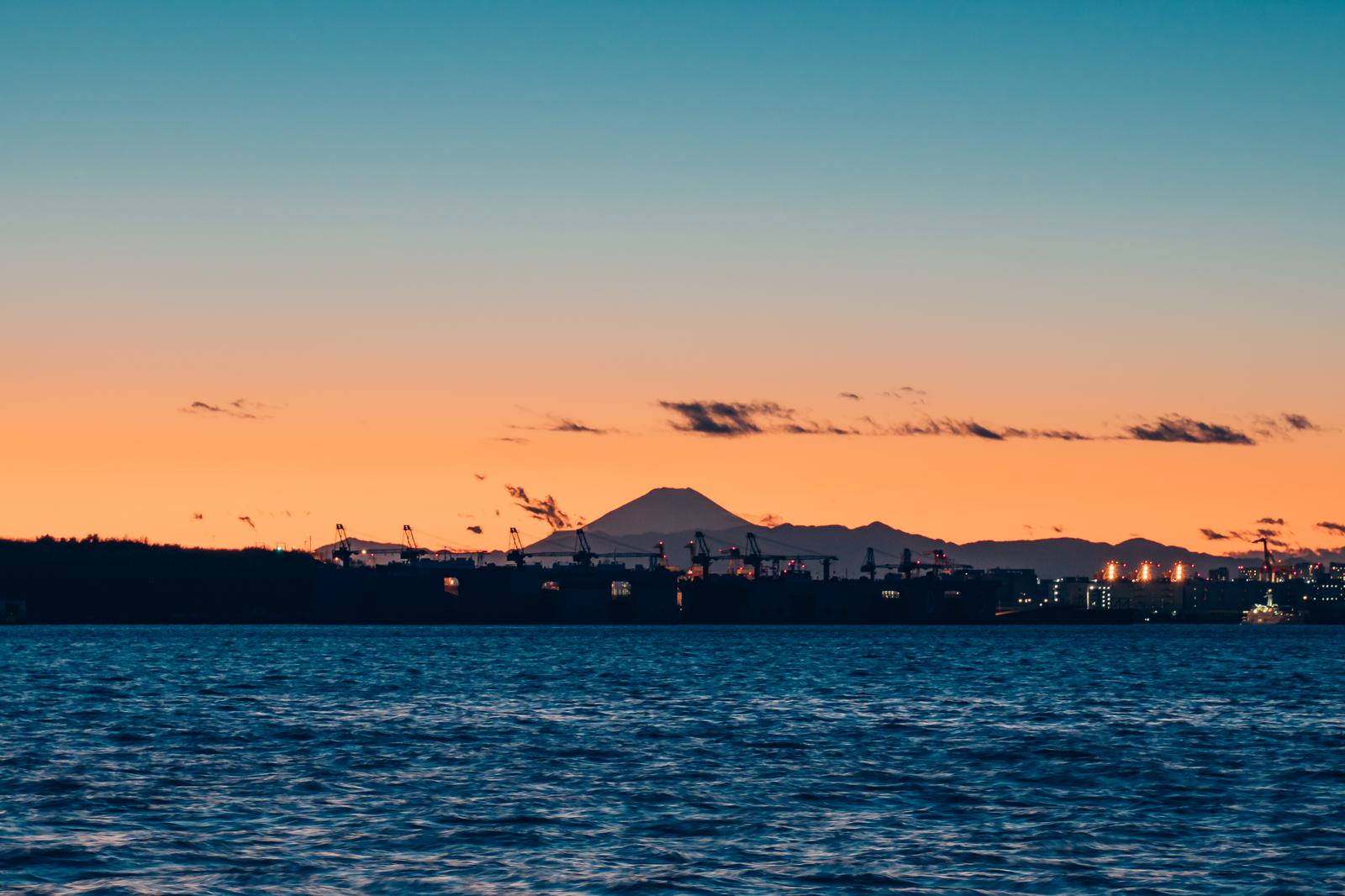 夕焼けに染まる空の下、東京湾越しに見える富士山と工業地帯のシルエット